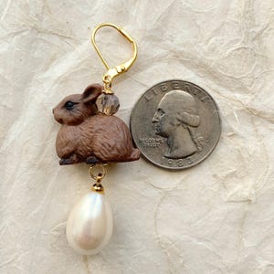 May include: A brown rabbit charm earring with a gold hook and a white pearl dangle. The earring is shown next to a US quarter for size comparison.