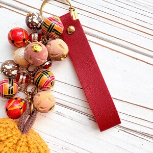 May include: A handmade keychain featuring a cluster of decorative beads in various colors and patterns, including red, brown, and gold. A red leather strap and gold-tone hardware complete the design. The keychain is attached to a gold ring.