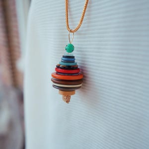 Puede incluir: Un collar con colgante único con un diseño de botones apilados en varios colores, incluyendo rojo, naranja, azul y marrón. El colgante está suspendido de una cadena dorada. Una pieza de joyería artesanal y artística.