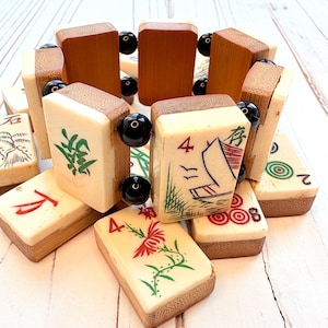 May include: A collection of vintage Mahjong tiles. The tiles feature ivory-colored faces with various symbols and numbers in red, green, and black. The tiles are framed with wood and connected with black beads. The game pieces are arranged on a white surface.