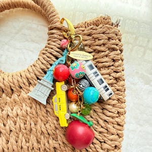 May include: A gold-toned metal keychain featuring charms of the Statue of Liberty, a yellow N.Y.C. taxi, a subway car, and a red apple, accented with colorful beads and a gold oval tag reading lenora dame MADE IN THE USA, attached to a woven bag.