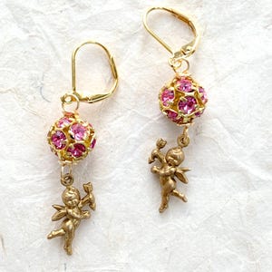 May include: Gold dangle earrings with a pink rhinestone ball and a gold cherub charm.