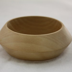 May include: A light-colored wooden bowl with a unique, geometric design. The bowl has a wide, open top and a gently sloping, angled exterior. The wood grain is visible, adding to the natural aesthetic. The bowl is likely used for decorative or functional purposes.