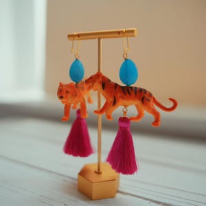 Op de afbeelding: Een paar unieke oorbellen met oranje tijgerfiguren met zwarte strepen, turquoise blauwe kralen en fuchsia kwastjes. De oorbellen worden gepresenteerd op een goudkleurige standaard, die hun speelse en artistieke ontwerp laat zien.