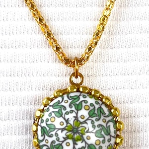 May include: Gold-toned necklace featuring an oval pendant with a white background and a green floral design. The pendant is framed with gold beads and hangs from a gold chain. A delicate piece of jewelry.
