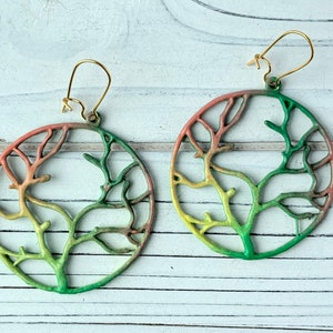 May include: A pair of circular earrings with a tree branch design. The earrings are made of metal and are painted in shades of green, yellow, and pink.