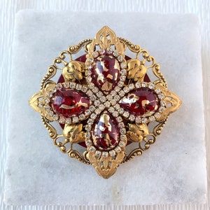Peut inclure: Une broche dorée avec un motif rouge et or. La broche présente quatre pierres rouges et or serties dans un cadre doré avec un motif décoratif.