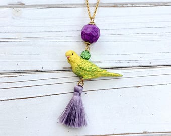 Puede incluir: Un collar de cadena dorada con una cuenta facetada morada, una cuenta verde y un dije de pájaro amarillo y verde. El collar termina con una borla morada.