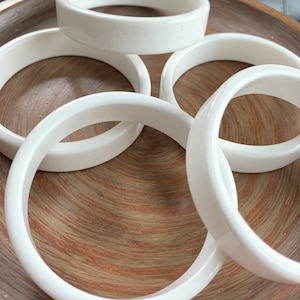 May include: A collection of white bangles, displayed on a wooden surface. The bangles are smooth and glossy, with a rounded shape. The image showcases the jewelry's clean design and bright white color.