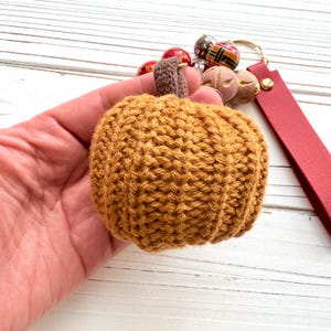 May include: A handmade, knitted pumpkin keychain in shades of orange and brown. The pumpkin has a textured appearance and a brown stem. The keychain includes a red leather strap, gold-colored hardware, and decorative beads.
