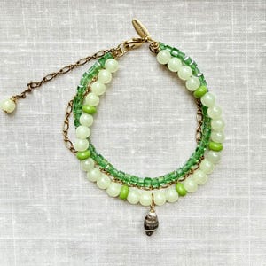 May include: A multi-strand bracelet featuring a gold-tone chain, translucent light green beads, and opaque pale green beads. The design includes a small metallic charm with the text 'lenora dame' and a single dark, textured, teardrop-shaped pendant.