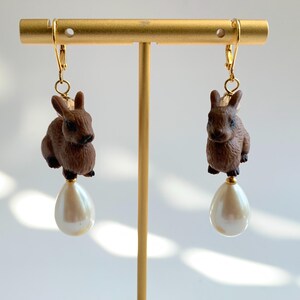 May include: A pair of gold dangle earrings featuring a brown rabbit charm and a white pearl drop.