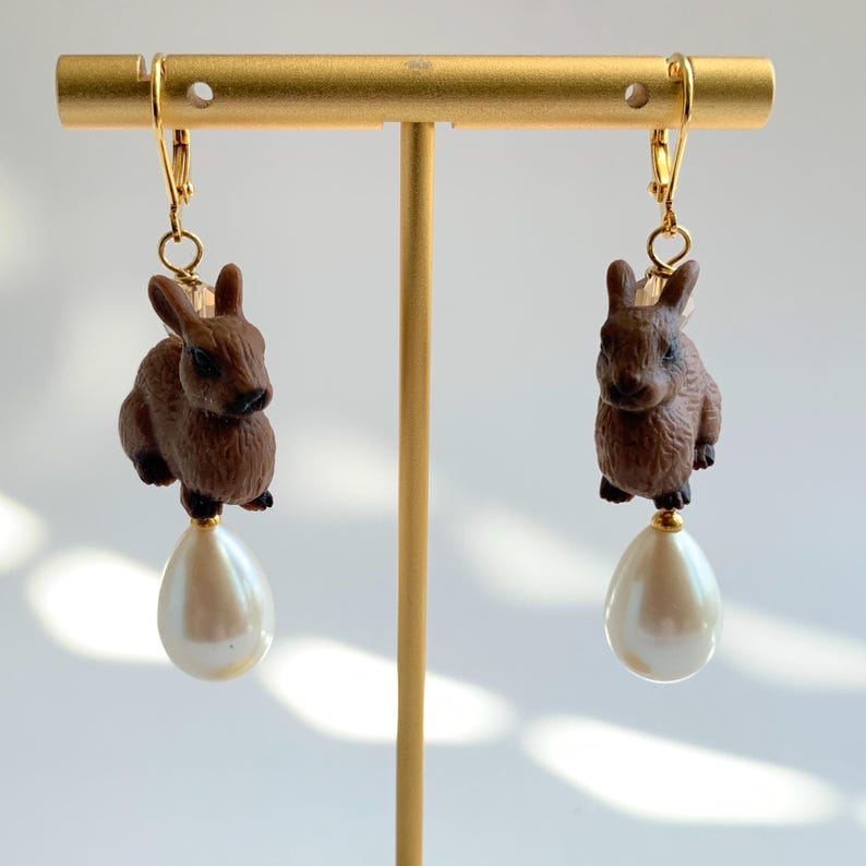 May include: A pair of gold dangle earrings featuring a brown rabbit charm and a white pearl drop.