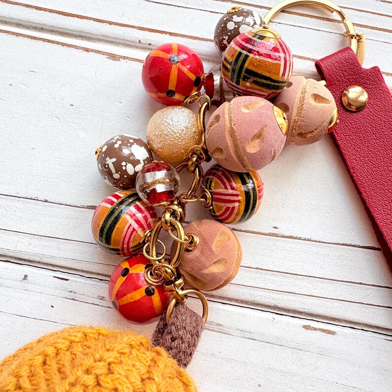 May include: A decorative keychain with a red leather strap and gold-tone hardware. The keychain features a cluster of colorful, patterned beads in various shapes and sizes, including red, brown, and pink spheres.