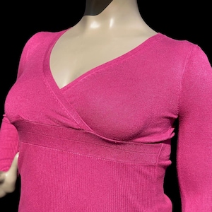 May include: A magenta sweater with a V-neck and long sleeves. The wrap-style design is at the chest, and the lower part has a ribbed texture. The garment is displayed on a mannequin against a black background.