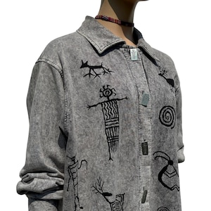 Könnte beinhalten: Graue Jeansjacke mit schwarzen, gestickten indianischen Petroglyphen-Designs. Die Jacke hat einen Kragen, lange Ärmel und Metallknöpfe. Die Jacke wird auf einer Schaufensterpuppe präsentiert.