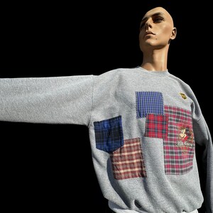 Peut inclure: Un sweat-shirt gris avec un motif patchwork composé de tissus à carreaux de différentes couleurs. Le sweat-shirt a un petit logo brodé sur la poitrine.