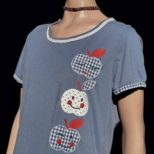 Puede incluir: Una camiseta azul con un cuello de encaje blanco y tres manzanas bordadas con caras sonrientes. Las manzanas son de cuadros rojos, blancos y azules.