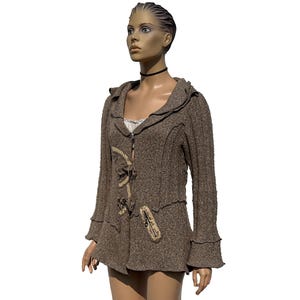 Peut inclure: Veste à capuche marron avec des accents décoratifs. La veste présente un design en tricot texturé, une fermeture éclair et des ornements uniques. Les manches ont un design évasé. La veste est présentée sur un mannequin.