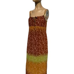 Op de afbeelding: Een lange, bruine, oranje en groene maxi-jurk met een bloemenprint en dunne bandjes. De jurk heeft een strik aan de bovenkant en een geplooide halslijn.