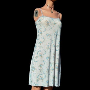 Puede incluir: Vestido lencero azul claro con estampado floral. El vestido presenta finos tirantes espagueti y un dobladillo a la altura de la rodilla. El diseño floral incluye flores azules y amarillas.