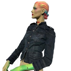 May include: A dark wash denim jacket with multiple pockets and button closures. The jacket is worn by a mannequin with colorful hair and makeup. The word "LOU" is visible on the mannequin's neck.