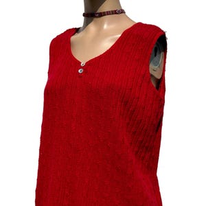 Puede incluir: Una camiseta roja sin mangas con un diseño de punto texturizado. La camiseta tiene un escote redondo y dos botones pequeños y redondos. La prenda se exhibe en un maniquí, mostrando su forma y estilo. También es visible un collar con estampado.