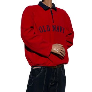 May include: A red fleece pullover with a navy blue collar and the text "OLD NAVY" in navy blue. The pullover has a quarter-zip closure and a drawstring at the waist. The model is wearing dark blue jeans.