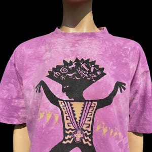 Op de afbeelding: Paarse tie-dye T-shirt met een zwart silhouet van een persoon met een hoofdtooi van symbolen en ontwerpen. De persoon danst met uitgestrekte armen.
