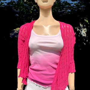 Puede incluir: Un top de tirantes de color rosa y blanco degradado con una chaqueta de punto de ganchillo rosa. La chaqueta tiene un corte holgado y un dobladillo ligeramente acampanado.