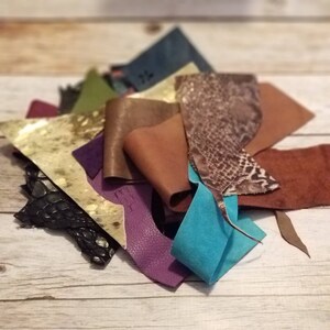 May include: A pile of assorted leather scraps in various colors and textures. The pieces include smooth, textured, and patterned leather in shades of brown, black, purple, green, and blue. The leather pieces are arranged on a light wood surface.