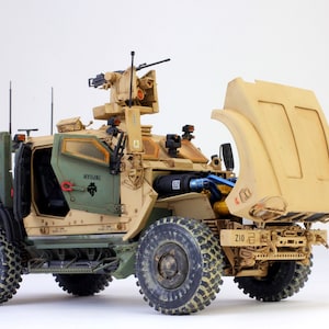 May include: A tan and green military vehicle with a raised gun turret and an open rear cargo area. The vehicle has a black and white "210" sticker on the back.