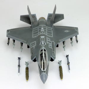 May include: A gray model F-35 fighter jet with detailed markings and attached missiles. Several model bombs are arranged around the jet. The aircraft has a clear cockpit and is set against a white background.