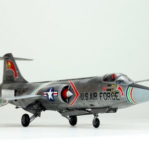 May include: A gray and silver Lockheed F-104 Starfighter model airplane with red, white, and blue markings. The model has a red star on the tail and the words "U.S. AIR FORCE" on the side. The model is sitting on a white surface.