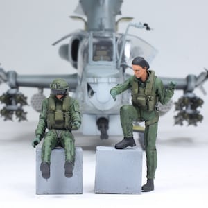 May include: Two military figures wearing green flight suits and helmets are sitting on concrete blocks in front of a model of a gray helicopter.