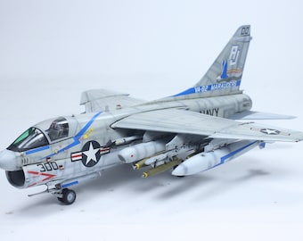 Pode incluir: Um modelo de avião A-7E Corsair II cinza e branco com um esquema de pintura azul e branco. O modelo está armado com bombas e foguetes e tem as marcas "VA-82 MARAUDERS" e "Navy" na fuselagem.