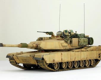 May include: A detailed model of a military tank in a tan color scheme. The tank features a long barrel, multiple turrets, and visible tracks. It has antennas and various storage compartments, showcasing a realistic design.