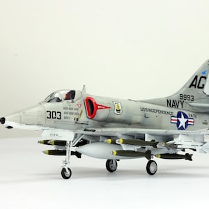 Puede incluir: Maqueta detallada de un avión A-4 Skyhawk gris claro, con el número 303 en el lateral. El modelo tiene una toma de aire roja y está equipado con misiles y un depósito de combustible. La cola presenta las marcas "AC" y "NAVY".