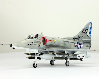 May include: A detailed model of a light gray A-4 Skyhawk aircraft, with the number 303 on the side. The model has a red air intake and is equipped with missiles and a fuel tank. The tail features the markings "AC" and "NAVY".