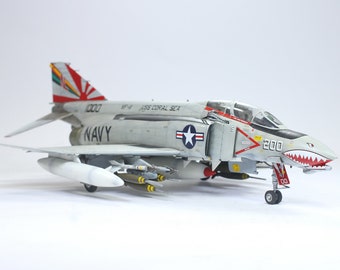 Pode incluir: Um modelo cinza e branco de um jato de combate McDonnell Douglas F-4 Phantom II com uma estrela vermelha, branca e azul no lado. O jato tem uma boca de tubarão pintada no nariz e o texto "VF-4 USS CORAL SEA 200" no lado. O jato está carregando várias bombas e mísseis.