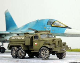 May include: A detailed model of an olive green fuel truck with the text "ОГНЕОПАСНО" and a red and white emblem. A light blue fighter jet is in the background. The truck has six wheels and a detailed front grill.