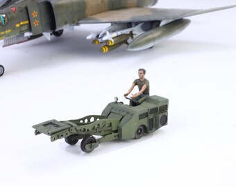 May include: A scale model of a military aircraft with a green and brown camouflage pattern. A small green vehicle with a figure is in the foreground. The model aircraft has missiles attached to the underside of the wings.
