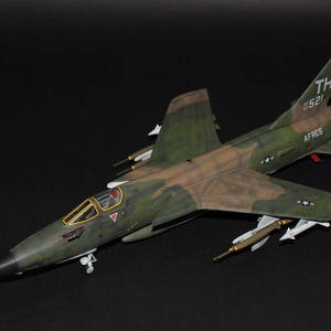 May include: A green and brown camouflage model airplane with the text "TH 0521" and "AFFRES" on the side. The model airplane has a long, pointed nose and two sets of wings.