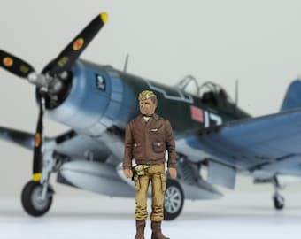 Może przedstawiać: Miniaturowa figurka pilota w brązowej skórzanej kurtce i khaki spodniach stoi przed niebiesko-szarym modelem samolotu. Pilot ma na sobie brązową skórzaną kurtkę i khaki spodnie. Samolot ma śmigło i czerwoną gwiazdę z białą obwódką na boku.