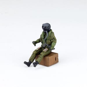 May include: A miniature figurine of a seated pilot in a green flight suit and black helmet. The pilot is seated on a small brown box. The figure has detailed features and is likely a model or collectible item.