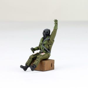May include: A miniature figurine of a pilot in a green flight suit, seated on a small brown box. The pilot is wearing a black helmet and has one arm raised. The figurine is detailed with realistic features and accessories.