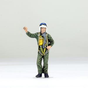 May include: A miniature figurine of a pilot wearing an olive green flight suit, a white helmet with blue goggles, and a yellow harness with silver buckles. The pilot has one arm raised and is equipped with a grey oxygen mask hose attached to the chest.