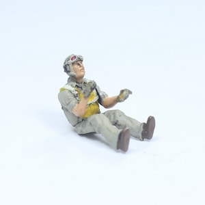 May include: A miniature figurine of a seated pilot, wearing a helmet with goggles, a yellow vest, and a gray uniform. The pilot is looking upwards with his hands raised. The figurine is painted in muted tones and sits on a white surface.