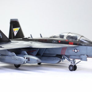 May include: A gray and black fighter jet model with red accents and a white nose cone. The model has two drop tanks and is sitting on a white surface. The jet has the number "360" on the side.