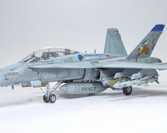 May include: A gray and white model of a fighter jet with the words "US NAVY" and "VIKINGS" on the side. The jet has a blue and white tail with a cartoon Viking on it. The jet is carrying several missiles and a fuel tank.